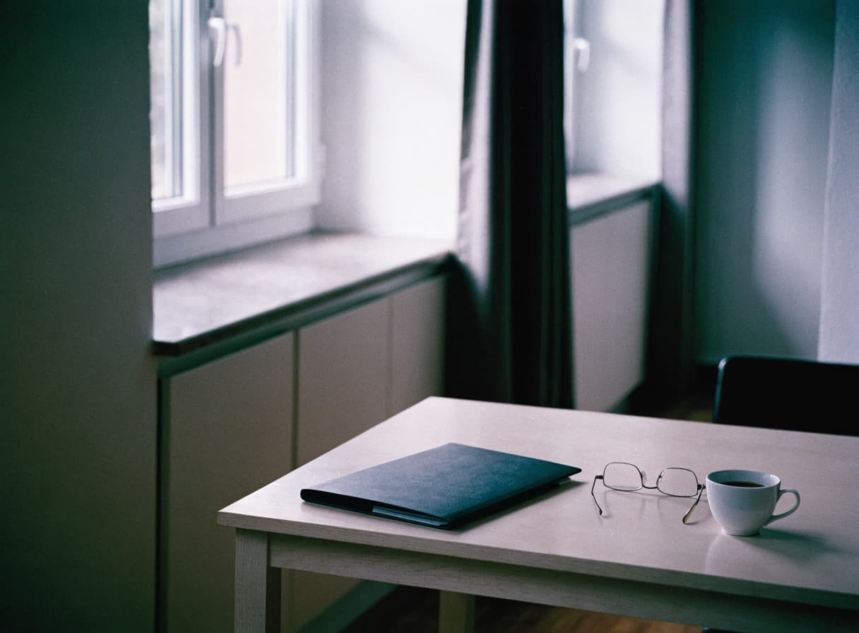 Geschlossene Mappe, Lesebrille und eine Tasse Kaffee auf einem Küchentisch am Fenster